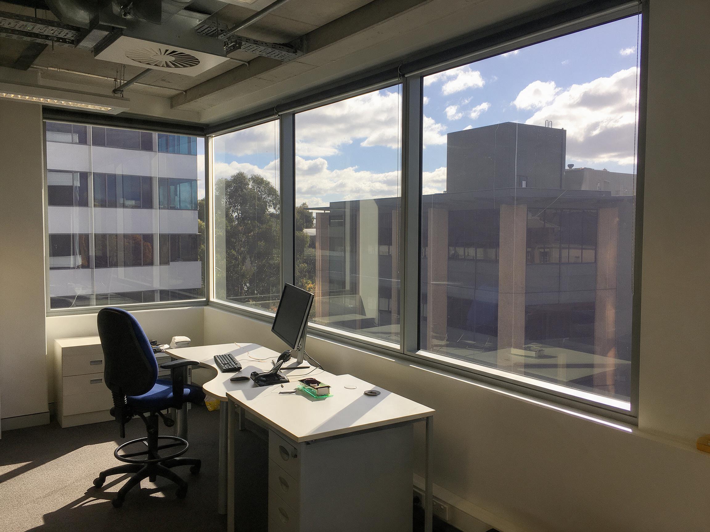 A working office beside a corner run of treated glazing, the desks and chairs evenly lit and the rooftops and sky outside still clearly visible