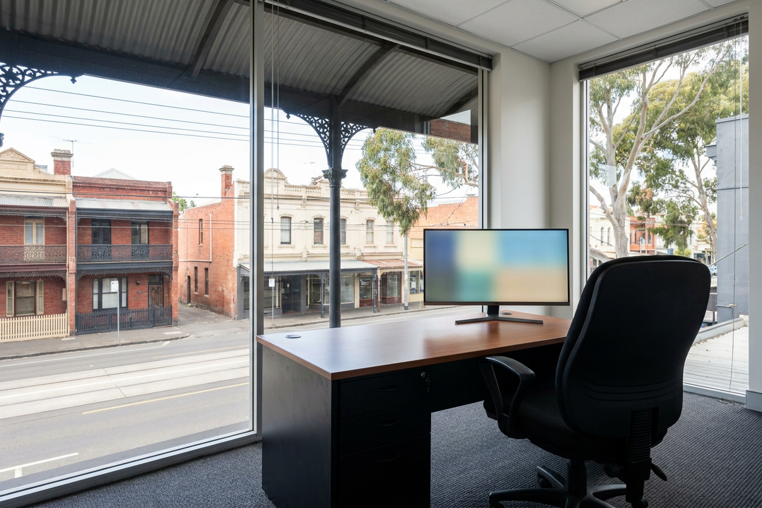 An office desk beside full height glazing onto a Melbourne street, the monitor screen legible against the daylight behind it