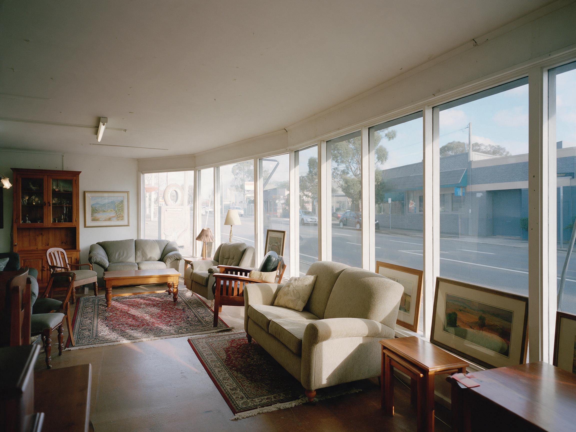 A furniture showroom behind a large shopfront window, upholstered lounges, timber and framed pictures standing close to the treated glass in full daylight